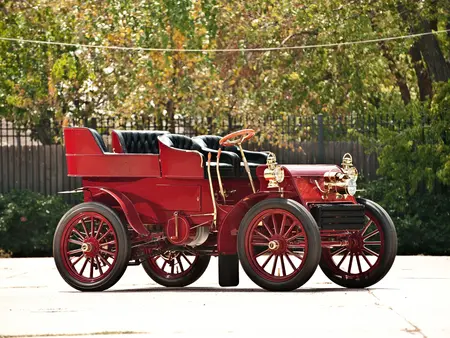 Red 1902 Packard Model F Vintage Convertible Car with red rims and black wheels, parked on concrete with a black fence and trees in the background