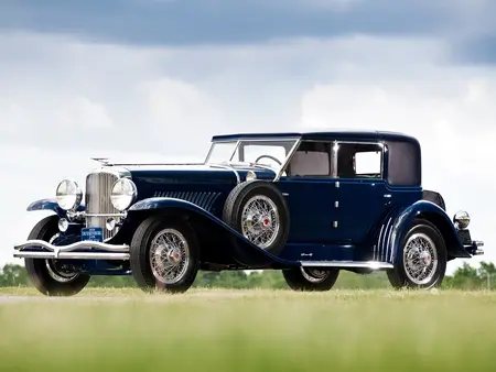 Black 1929 Duesenberg Model J with silver rims, parked on grass with clouds in the background