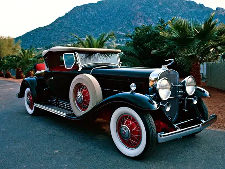 Black Vintage Cadillac Model 452 with red wheels with white accents, parked on concrete with palm trees and a mountain in the background