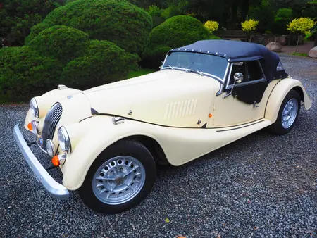 Cream colored 1930 Morgan plus 8 Vintage Car with black roof and grey rims, parked on asphalt with green bushes in the background