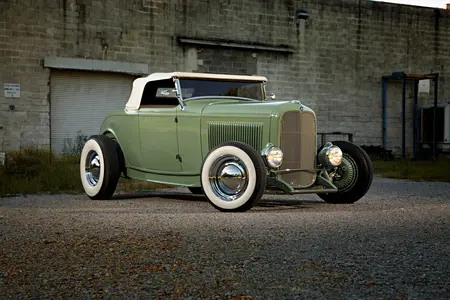 Off Green 1932 Ford Roadster with silver plate rims and white accent, parked on conrete outside a dirty garage