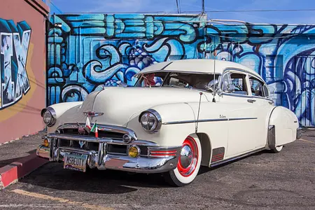 Cream Colored 1949 Chevrolet Fleetline with red plate rims and white accents, parked on concrete with blue graffiti on the background